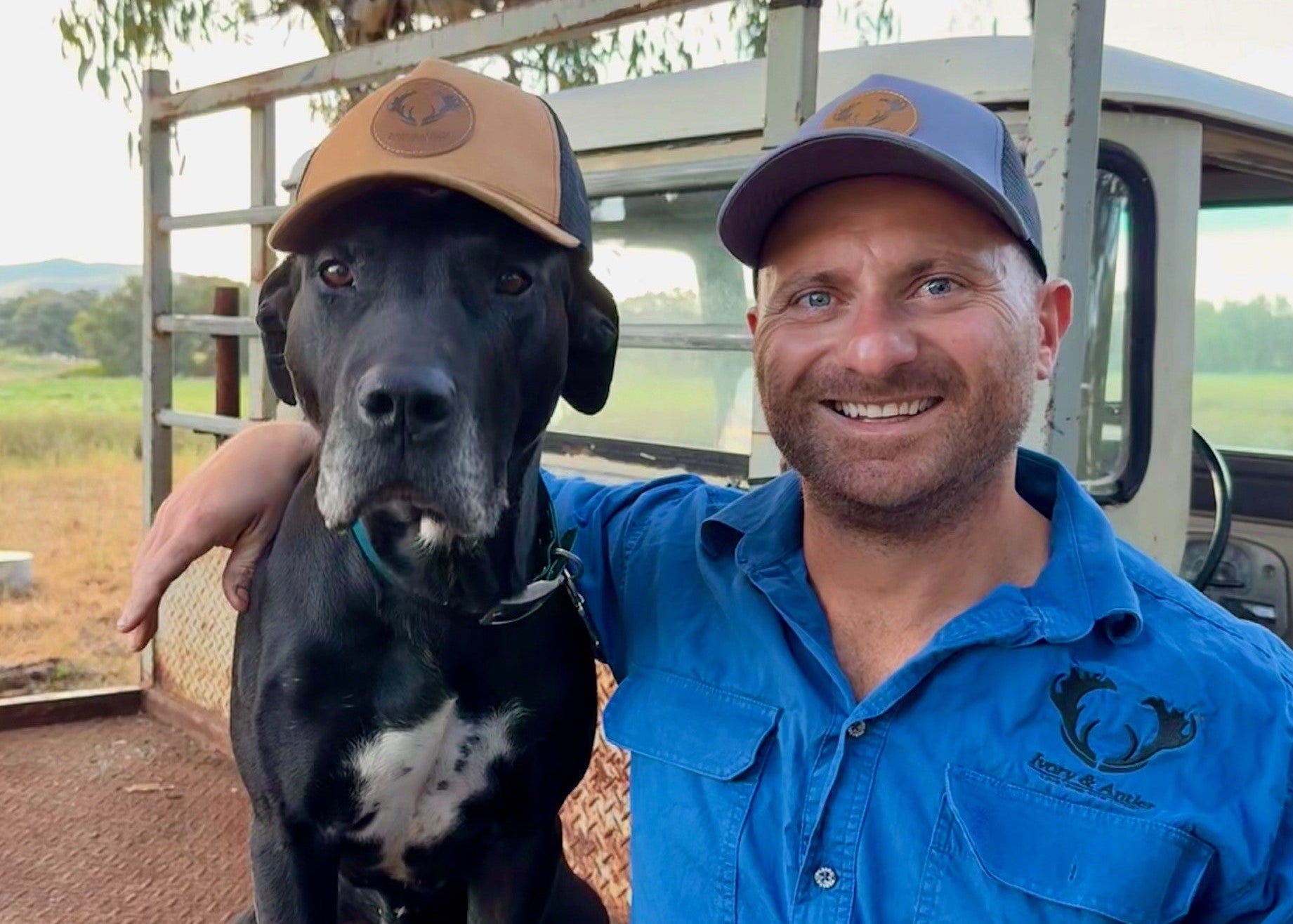Man in blue shirt and cap with a black dog on a truck bed outdoors