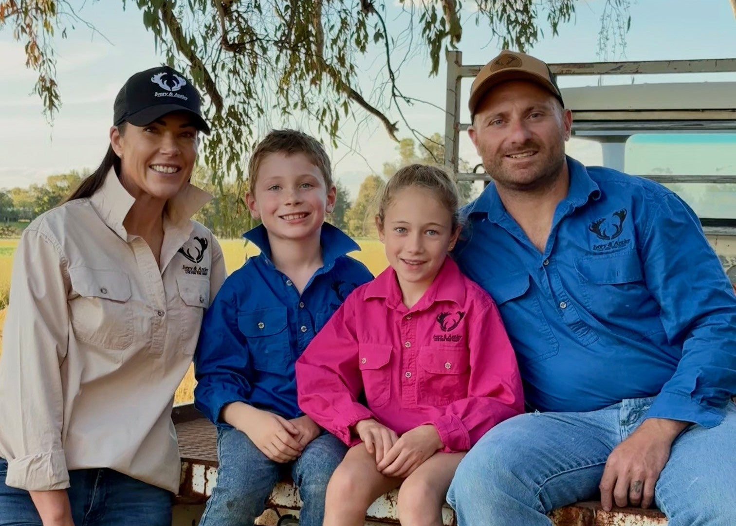 Family of four sitting on a truck bed outdoors with a tree and open field in the background.